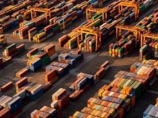 Aerial view: Hundreds of brightly colored shipping containers are stacked high at a busy port/terminal, with large yellow gantry cranes...