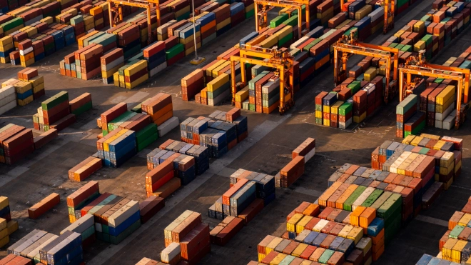 Aerial view: Hundreds of brightly colored shipping containers are stacked high at a busy port/terminal, with large yellow gantry cranes...